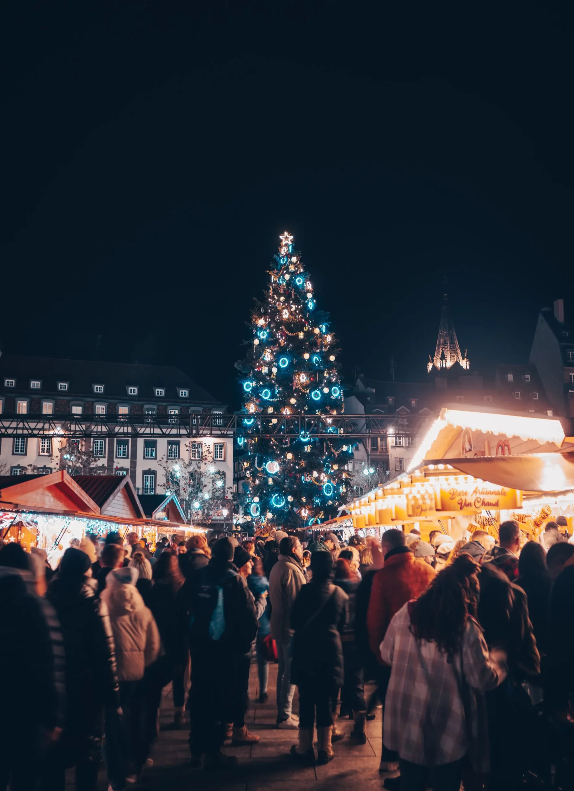Un affollato mercatino di Natale a Strasburgo di notte, con un maestoso albero di Natale illuminato da luci blu e bianche, circondato da chalet in legno accoglienti e visitatori in abiti invernali.