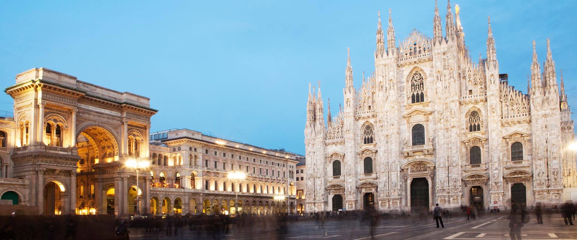 Vista notturna del Duomo di Milano illuminato, con la Galleria Vittorio Emanuele II a sinistra e persone che camminano nella piazza.