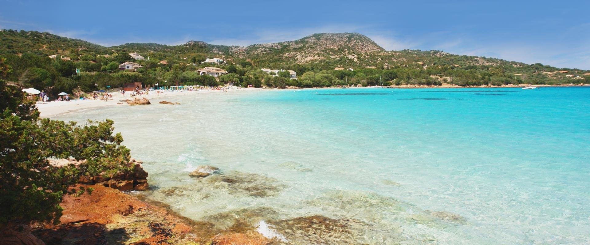 Vista di una spiaggia di sabbia bianca con acqua turchese cristallina in Sardegna, Italia, con rocce rossastre in primo piano e vegetazione verde sulle colline circostanti.